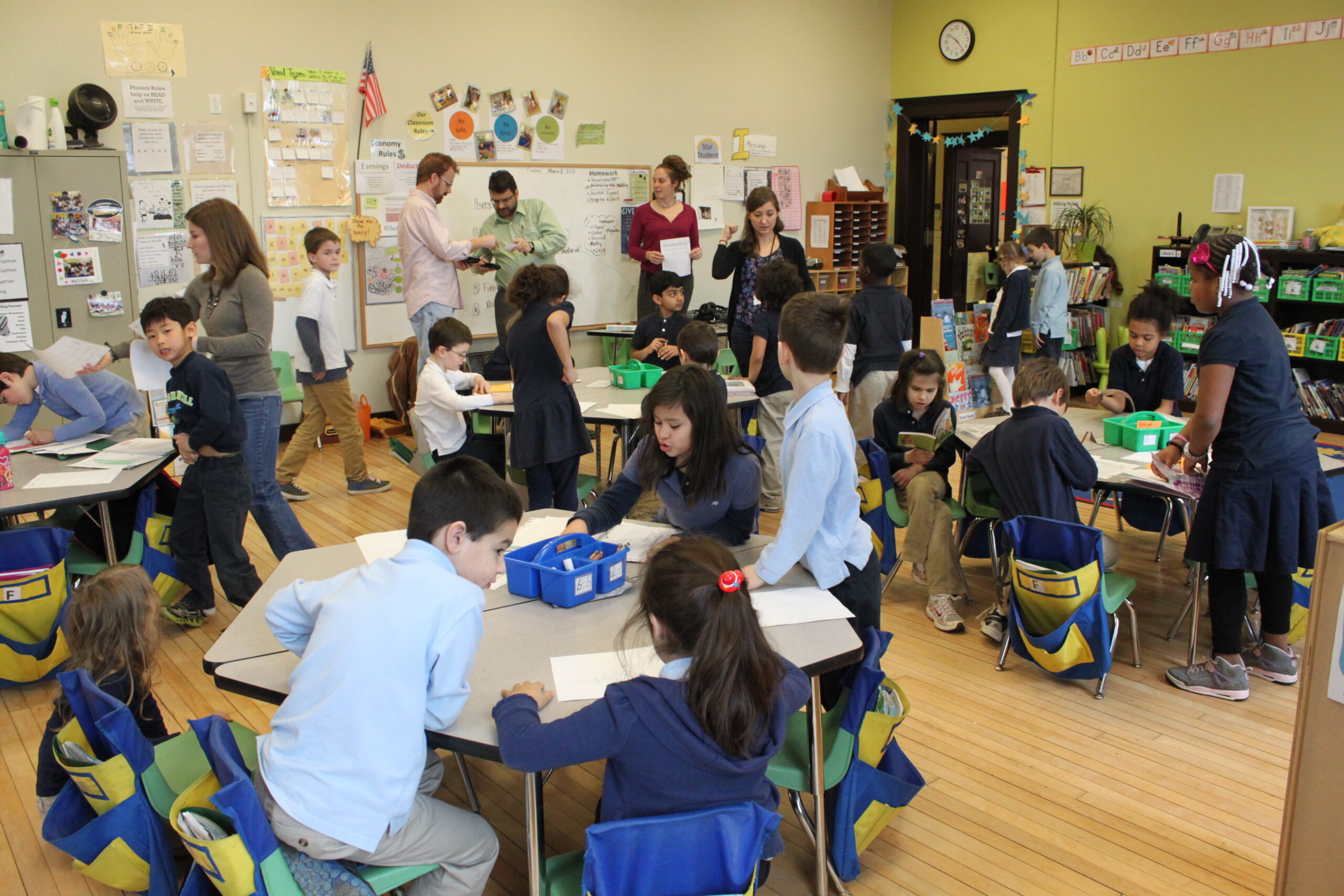 Eighteen youth, eleven sitting at classroom desks, seven standing around the desks, and five adults overseeing while standing in front of a whiteboard.