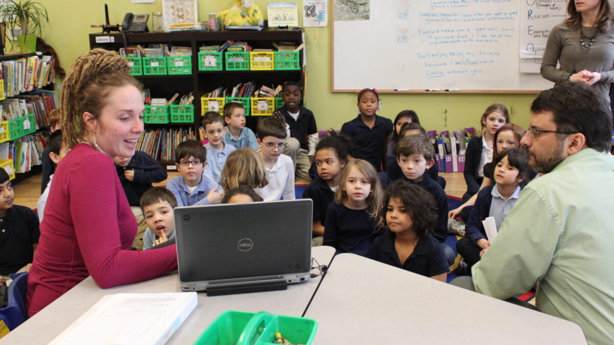 Twenty-two youth sitting on the floor, an adult standing in front of a whiteboard, and an adult sitting at a classroom desk, all looking at another adulting interacting with a laptop computer on a classroom desk