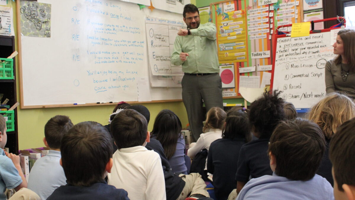 Twelve youth sitting on the floor and an adult standing next to a large posterboard looking at an adult gesturing with their hands in front of a whiteboard in a classroom