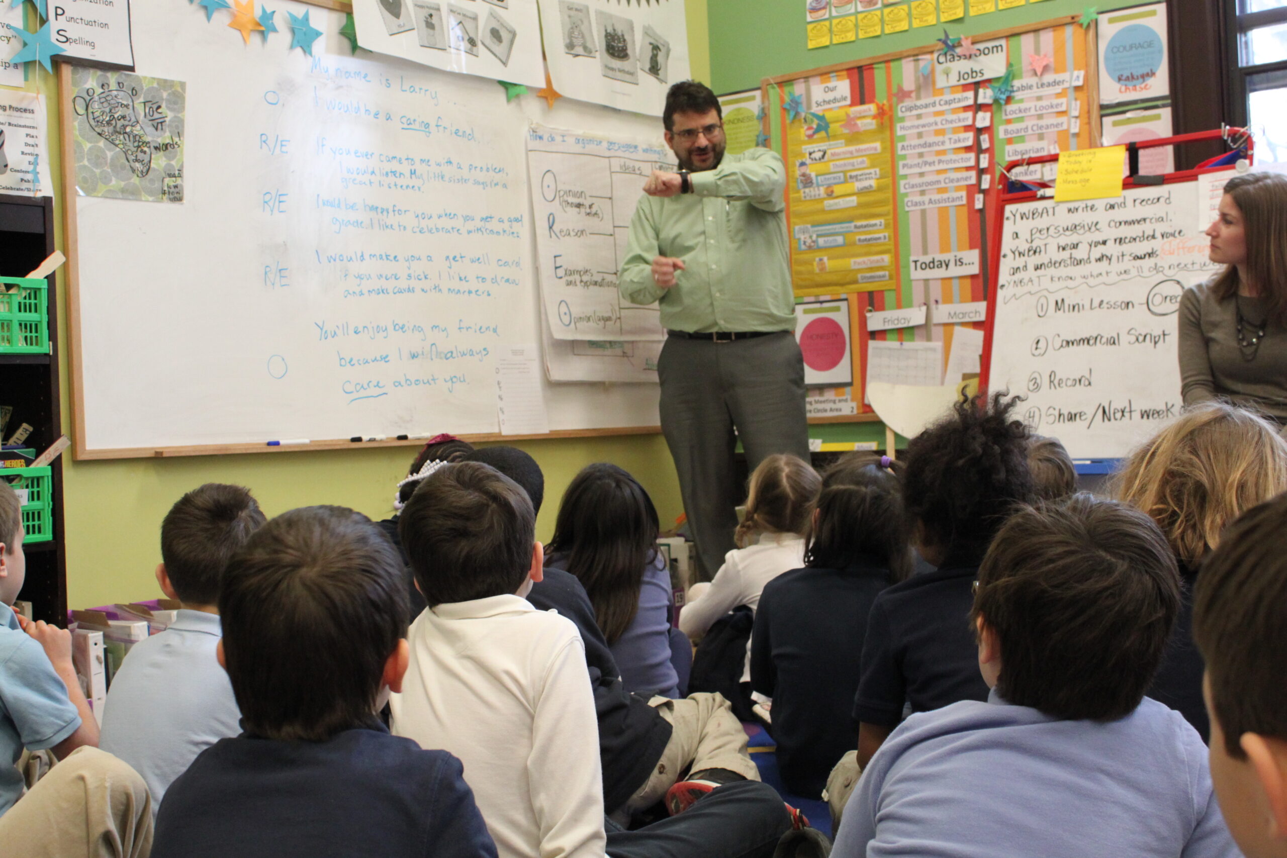 Twelve youth sitting on the floor and an adult standing next to a large posterboard looking at an adult gesturing with their hands in front of a whiteboard in a classroom