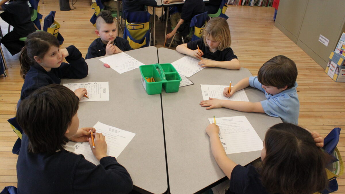 Seven youth writing on pieces of paper on a classroom desk