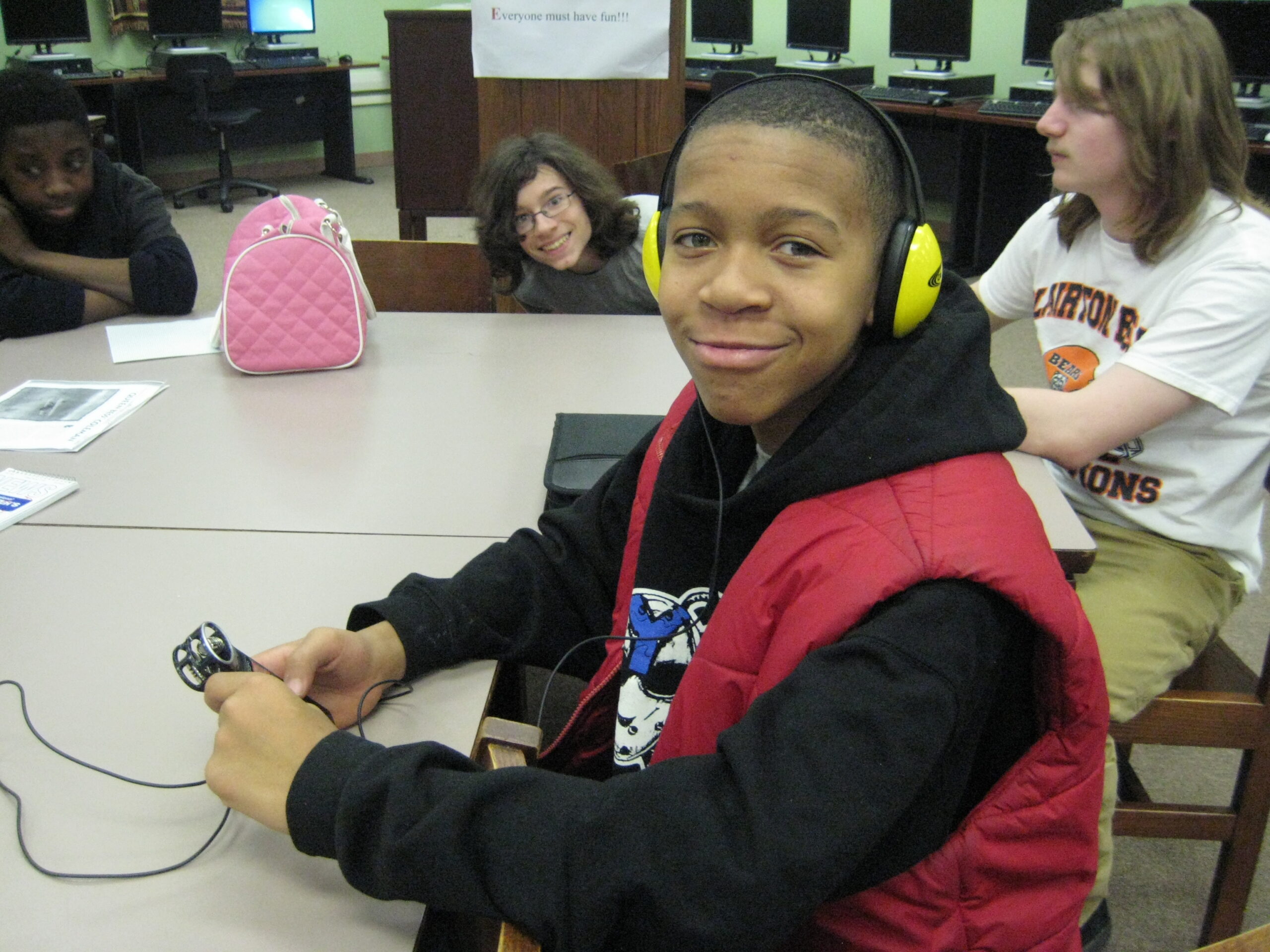 A youth wearing headphones holding a portable recorder and smiling at the camera in a classroom