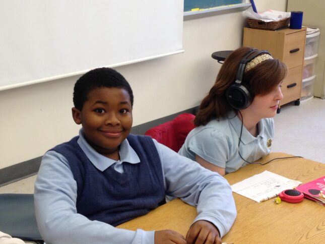A youth sitting and smiling at the camera and a youth wearing headphones sitting behind a piece of paper