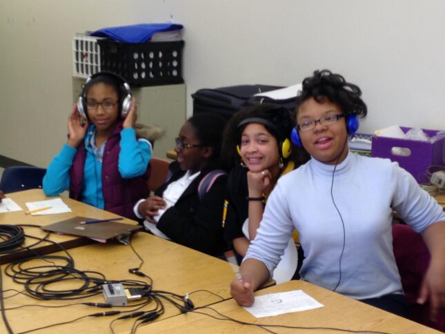Four youth wearing headphones connected to an audio splitter smiling at the camera and sitting at classroom desks