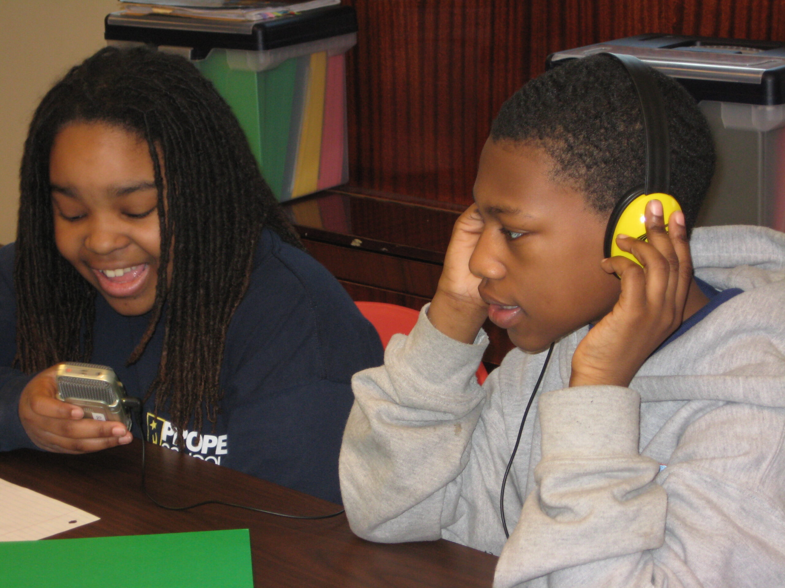 A youth wearing headphones and a youth smiling while holding a portable recorder, both sitting at a wooden table