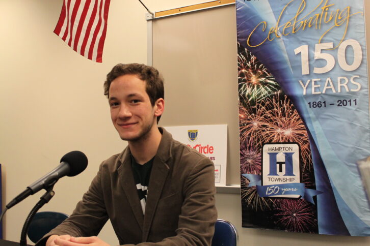 An adult sitting behind a microphone and in front of a banner celebrating 150 years of Hampton Township