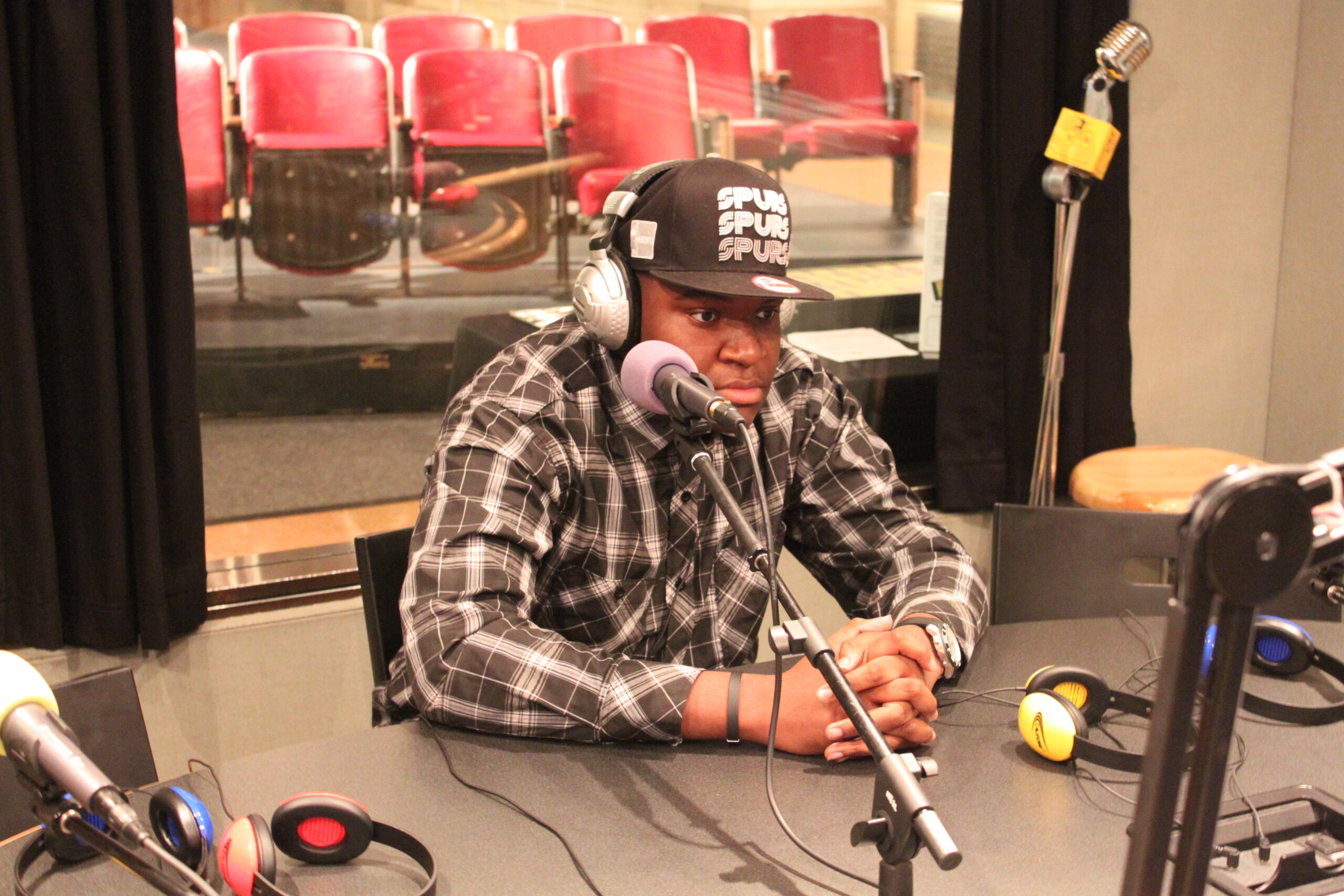 A teen wearing headphones and a ball cap sitting behind a microphone in a radio studio