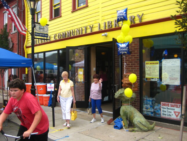 Two adults and a youth leaving a yellow building with brick walls and the text "community library" on the top, and a green statuette holding balloons out front.