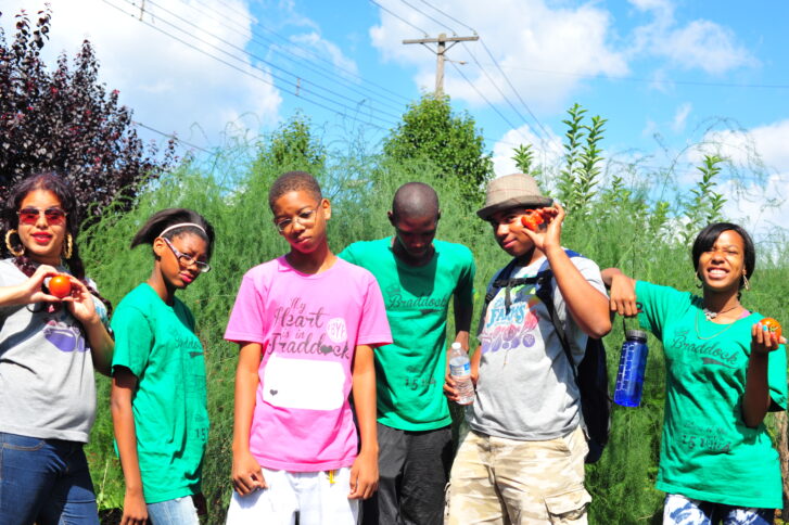 Five teens and an adult posing in front of large bushes under a blue, sparsely cloudy sky