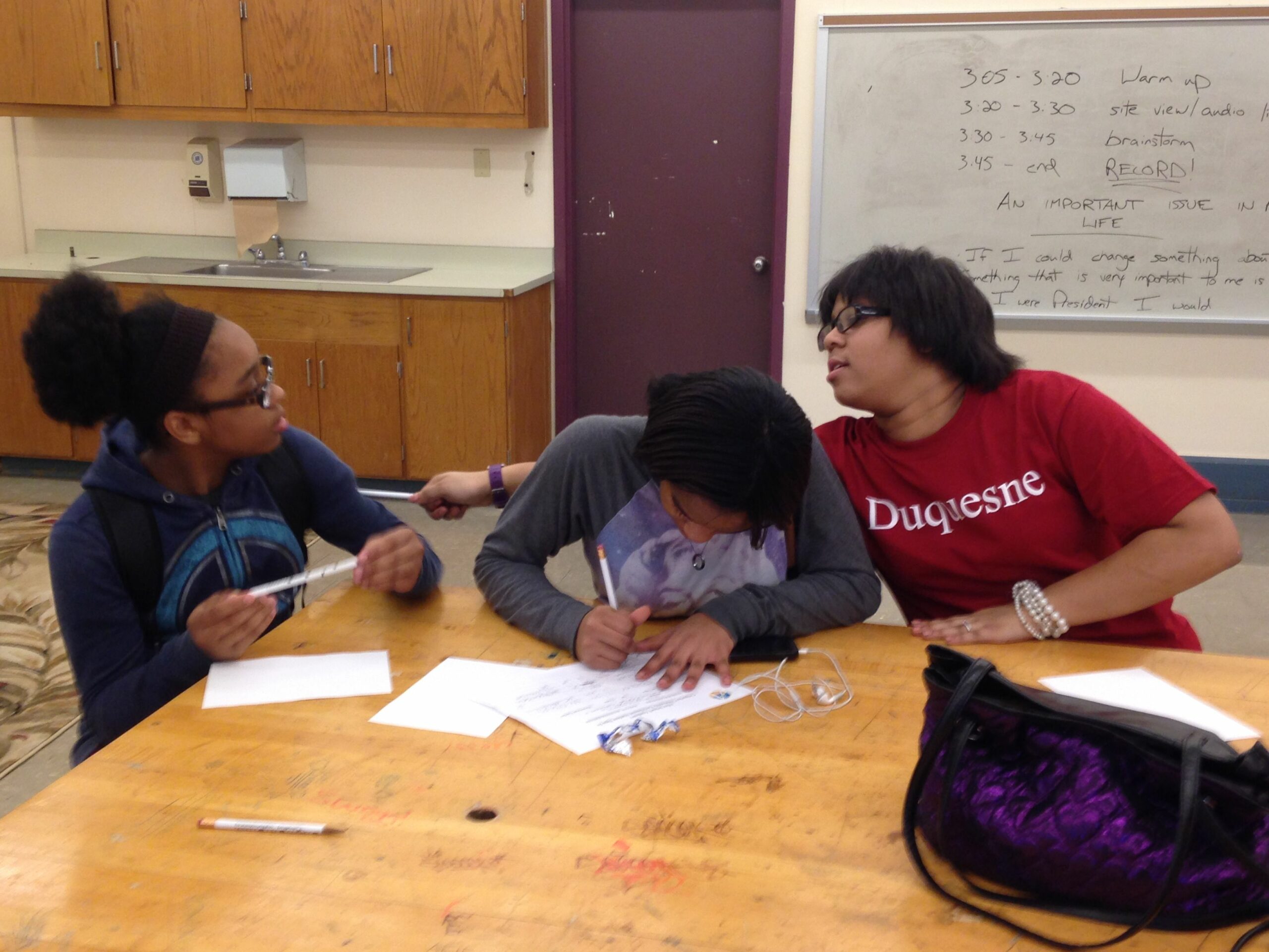 Two youth looking at each other over another youth's back writing on a piece of paper on a classroom table