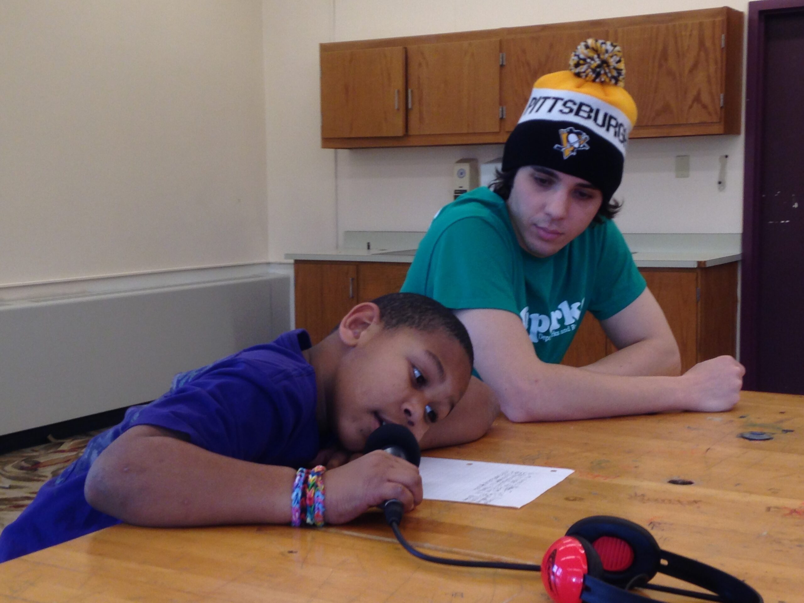 A youth looking down at another youth resting their head on a table while speaking into a microphone and looking at a piece of paper