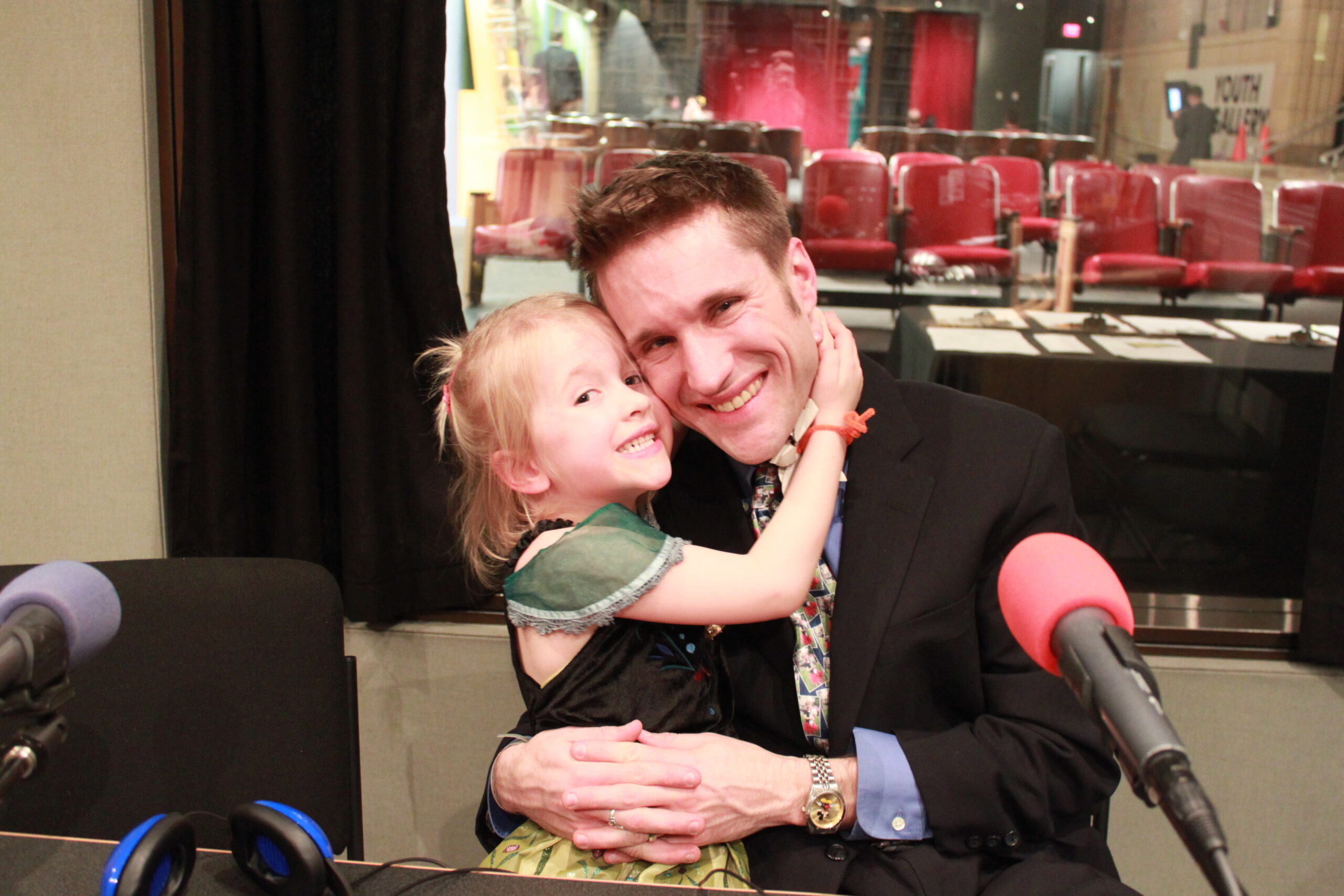 A youth and an adult holding each other while sitting behind a microphone in a radio studio
