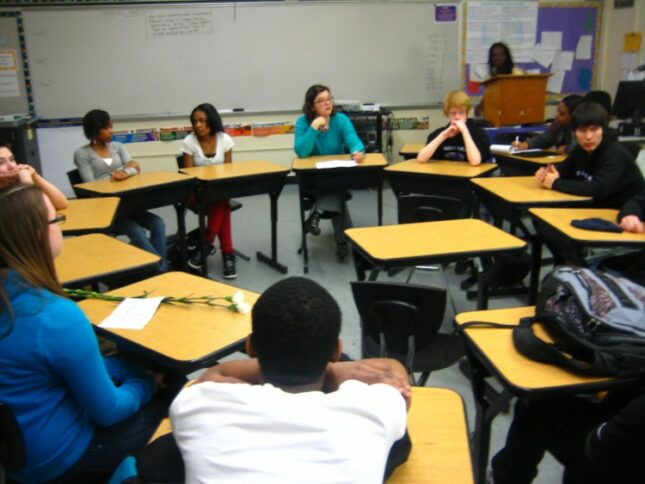 Eight youth sitting at desks in a circle in a classroom in front of a whiteboard