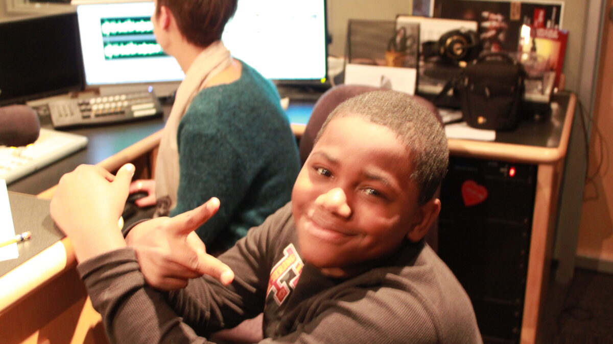 A youth smiling at the camera and pointing at an adult sitting behind computer screens in a radio studio