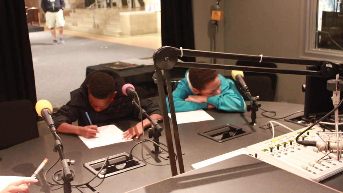 One youth writing on a piece of paper and one youth with their head down on their arms in a radio studio