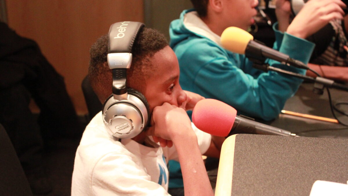 One youth sitting while wearing headphones and one youth sitting while speaking into a microphone in a radio studio