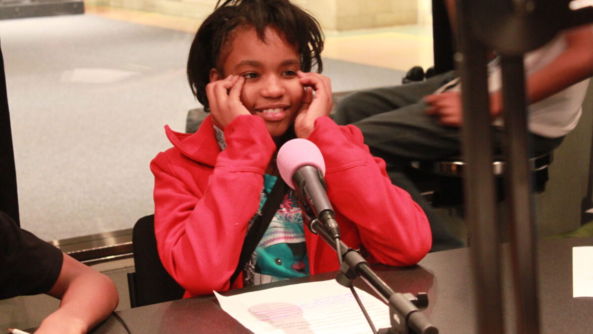 A youth holding their face in their hands while sitting behind a microphone and a piece of paper on a table in a radio studio