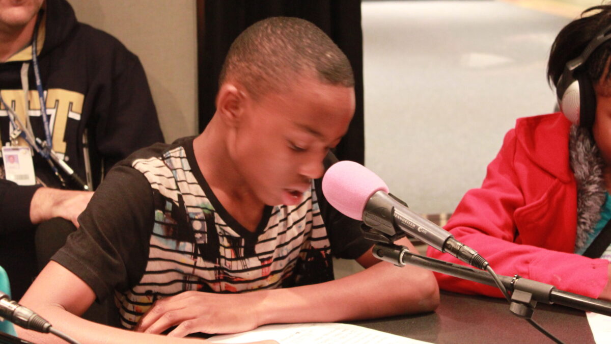 A youth speaking into a microphone and looking down at a piece of paper in a radio studio