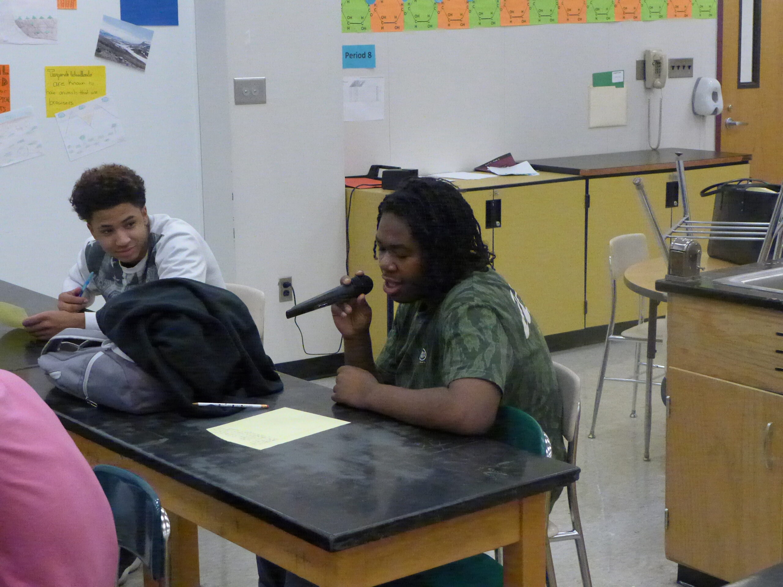 A youth looking at another youth holding and speaking into a microphone, both sitting at a classroom desk