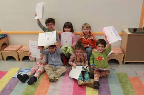 Seven children holding up drawings while sitting on a multicolored floor