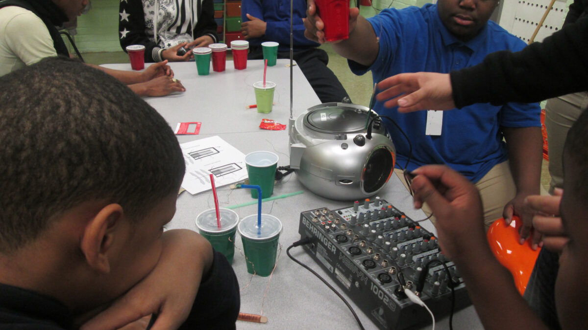 Five youth sitting around a cd player and a mixer on a white table in a classroom, one youth holding up a red solo cup