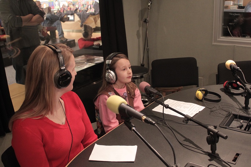An adult and a youth wearing headphones speaking into microphones in a radio studio