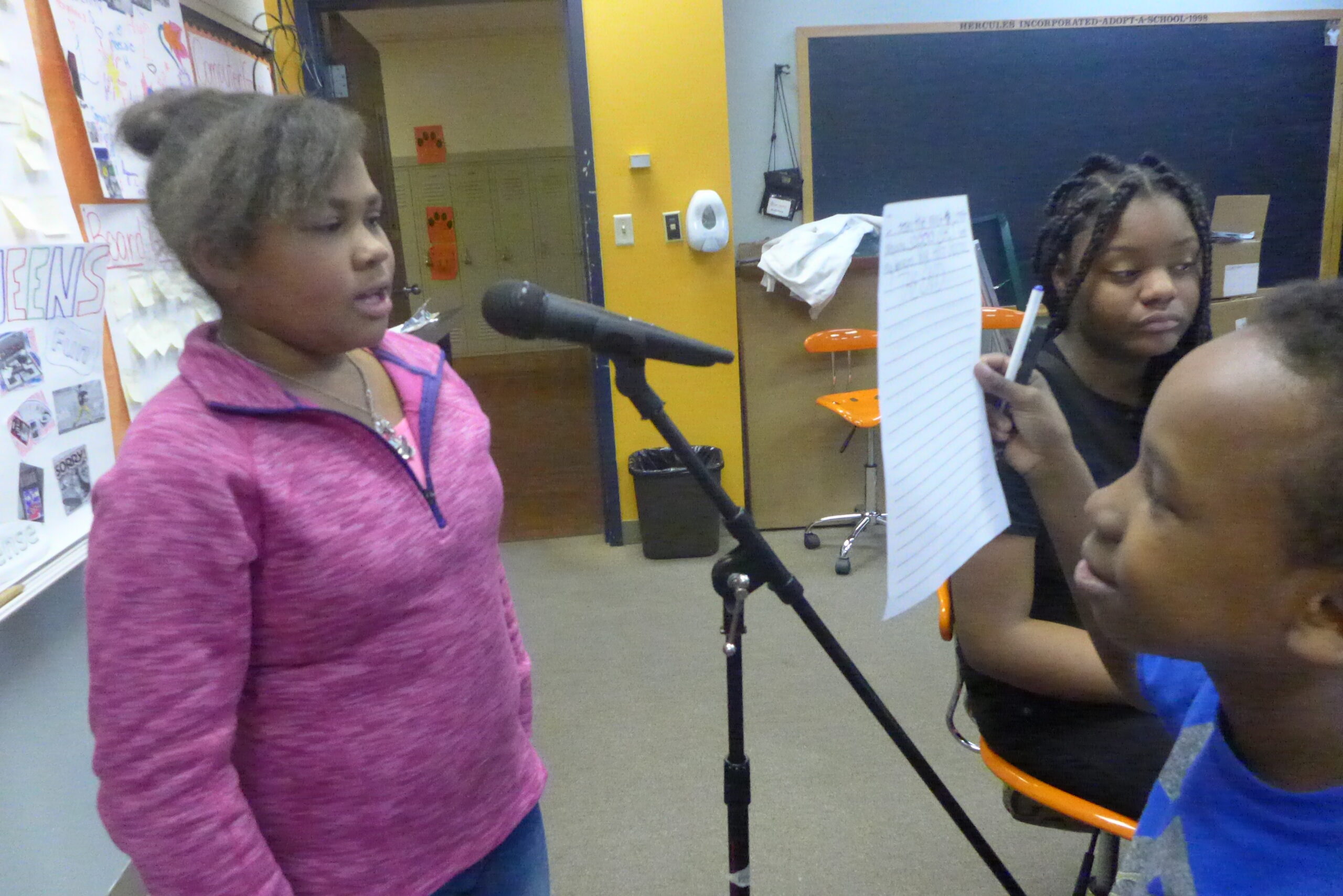 A youth holding up a piece of paper for another youth speaking into a microphone as another youth sits nearby in a classroom