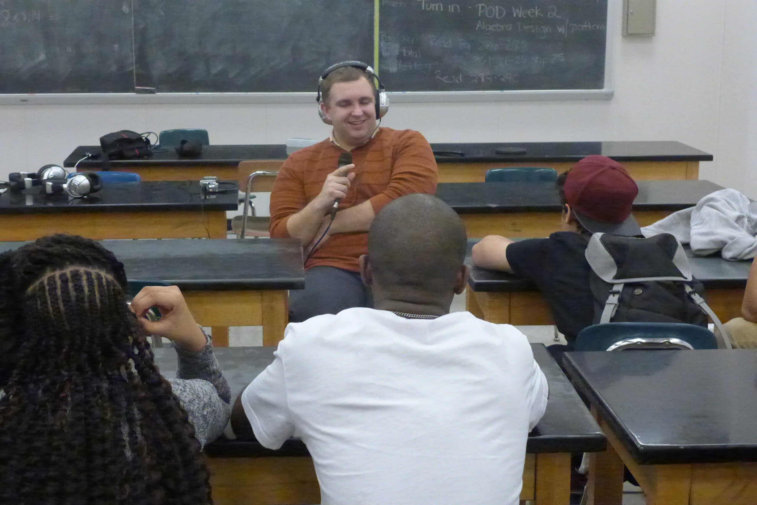 Three youth sitting at classroom desks listening to an adult wearing headphones holding and speaking into a microphone in a classroom