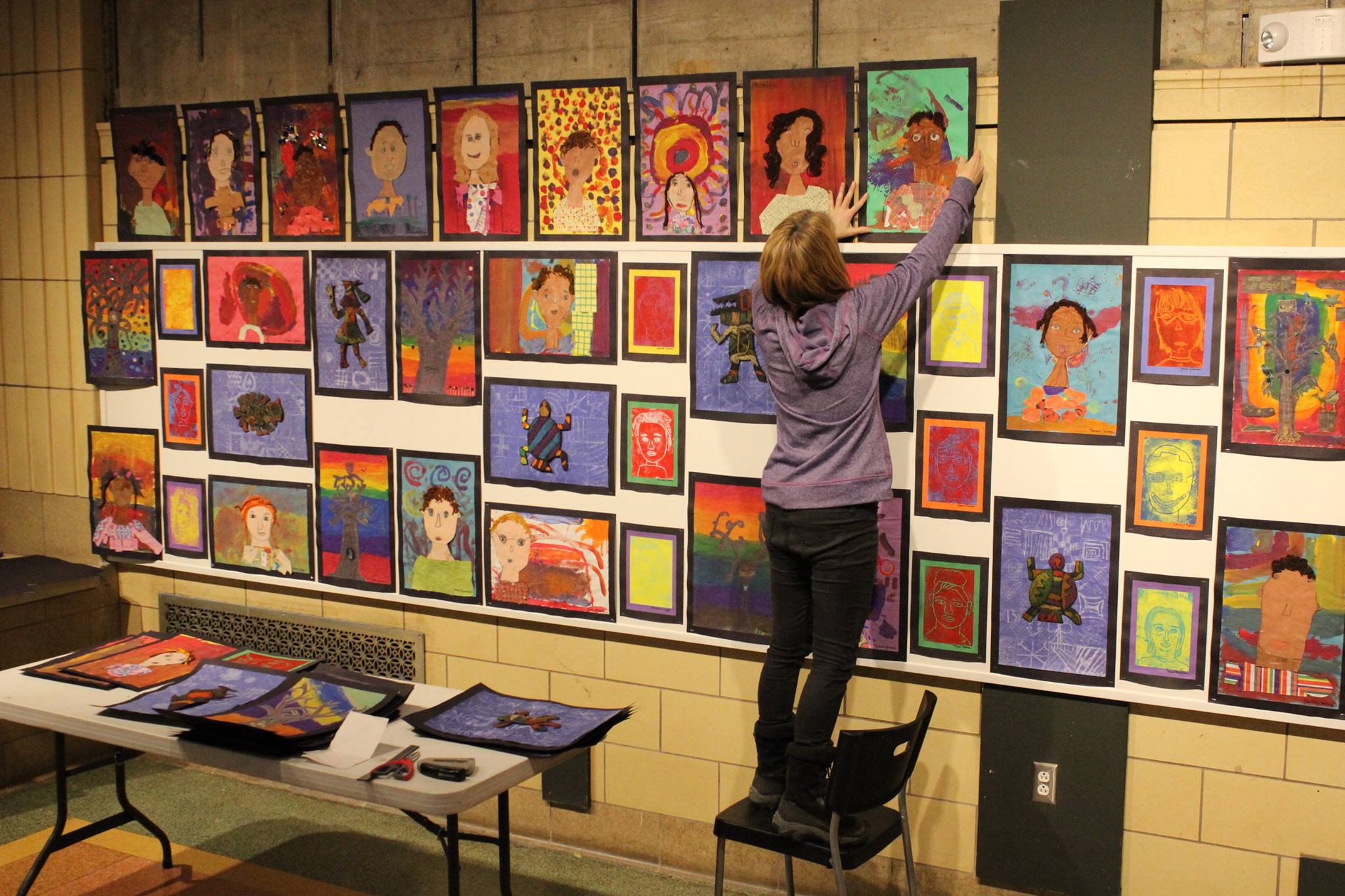An individual standing on a chair hanging up a painting on a wall covered with other paintings