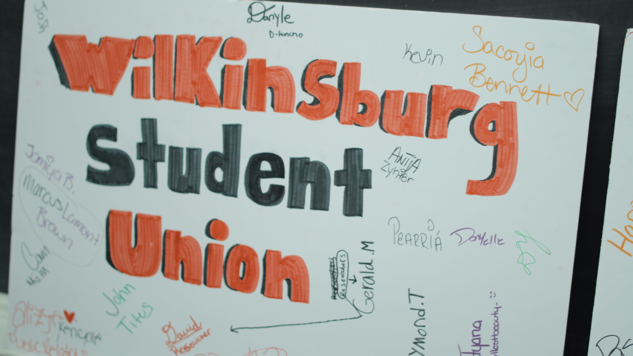 A poster board with the text "Wilkinsburg" in orange, "Student" in dark gray, and "Union" in orange surrounded by signatures