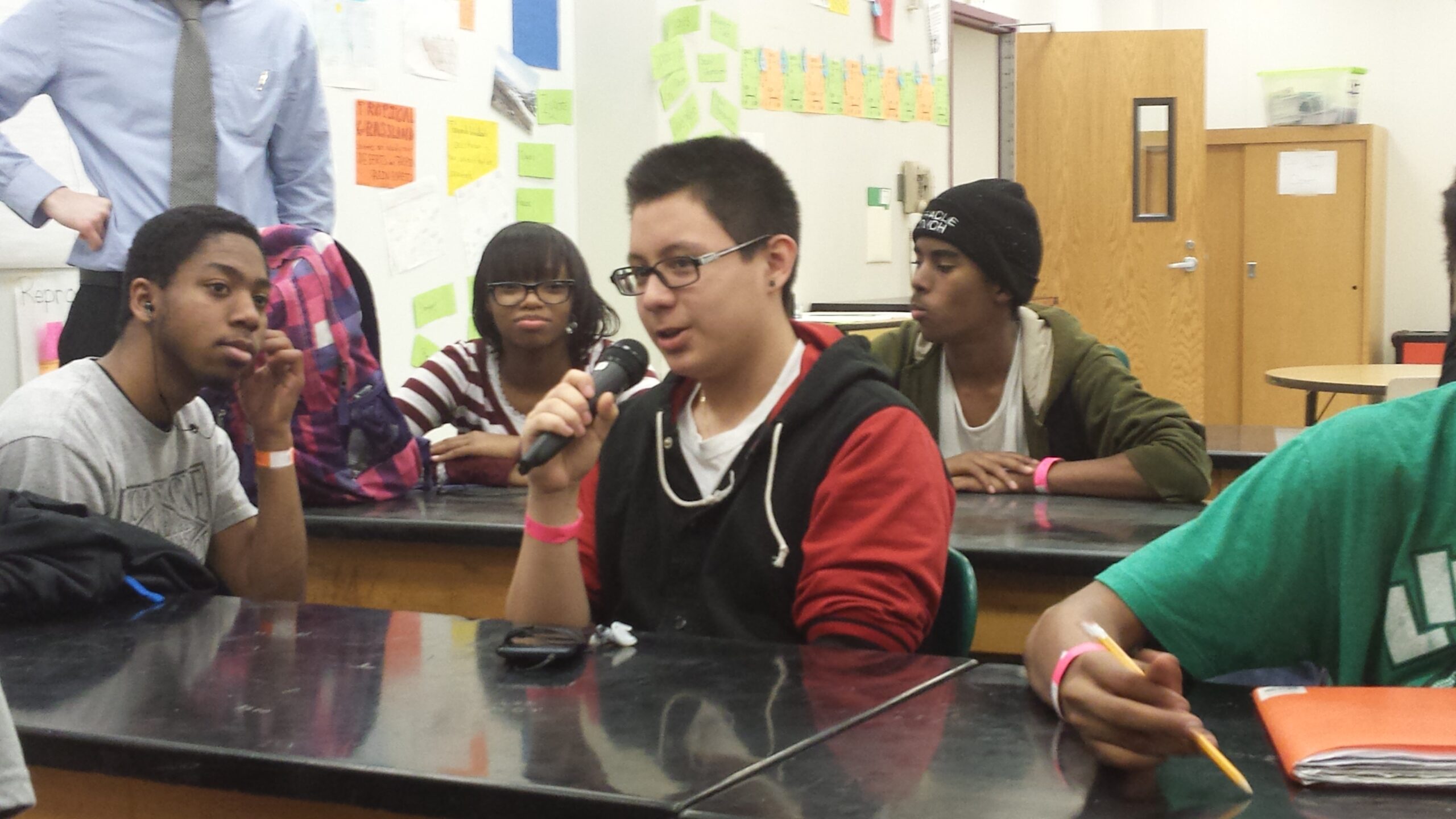 Four teens sitting at classroom desks, one holding and speaking into a microphone