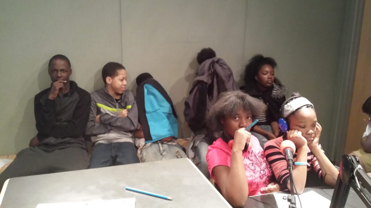 Five youth sitting in a radio studio, two of the youth wearing headphones sitting behind microphones