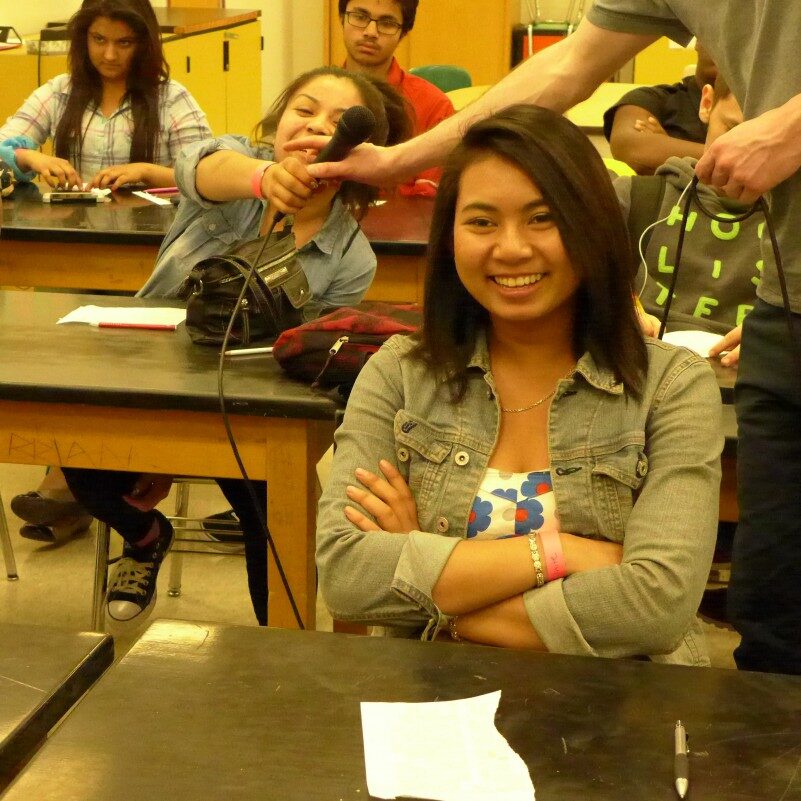 Two youth sitting at classroom desks, one smiling while crossing their arms, the other attempting to grab a microphone from an adult's hand