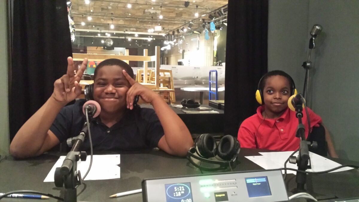Two youth wearing headphones sitting behind microphones in a radio studio, one flashing two peace signs