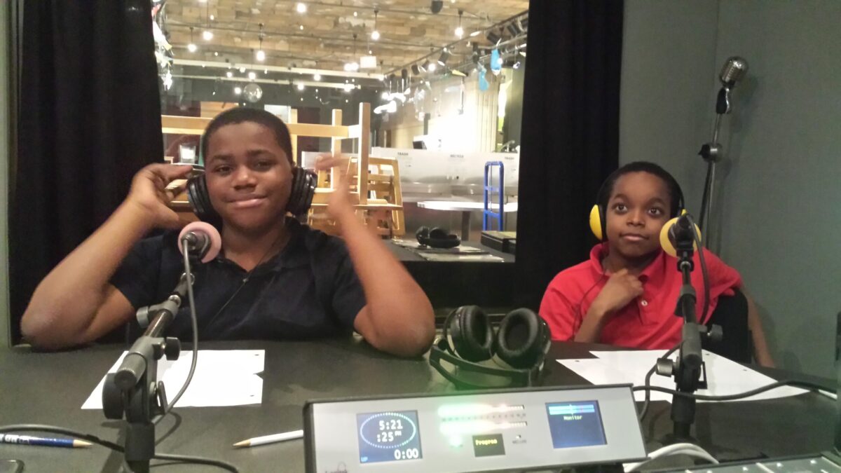 Two youth wearing headphones sitting behind microphones in a radio studio.