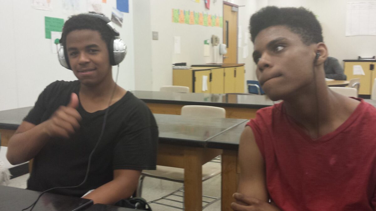 Two teens sitting in a classroom, one wearing headphones and giving a thumbs up to the camera