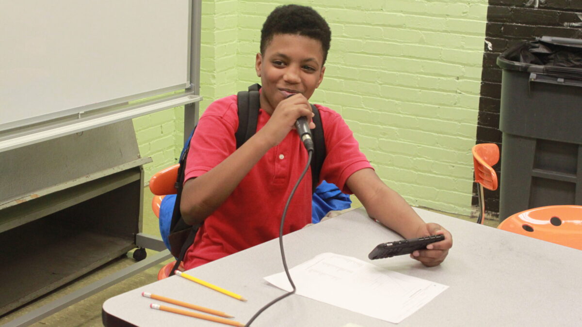 A youth holding and speaking into a microphone sitting in a classroom