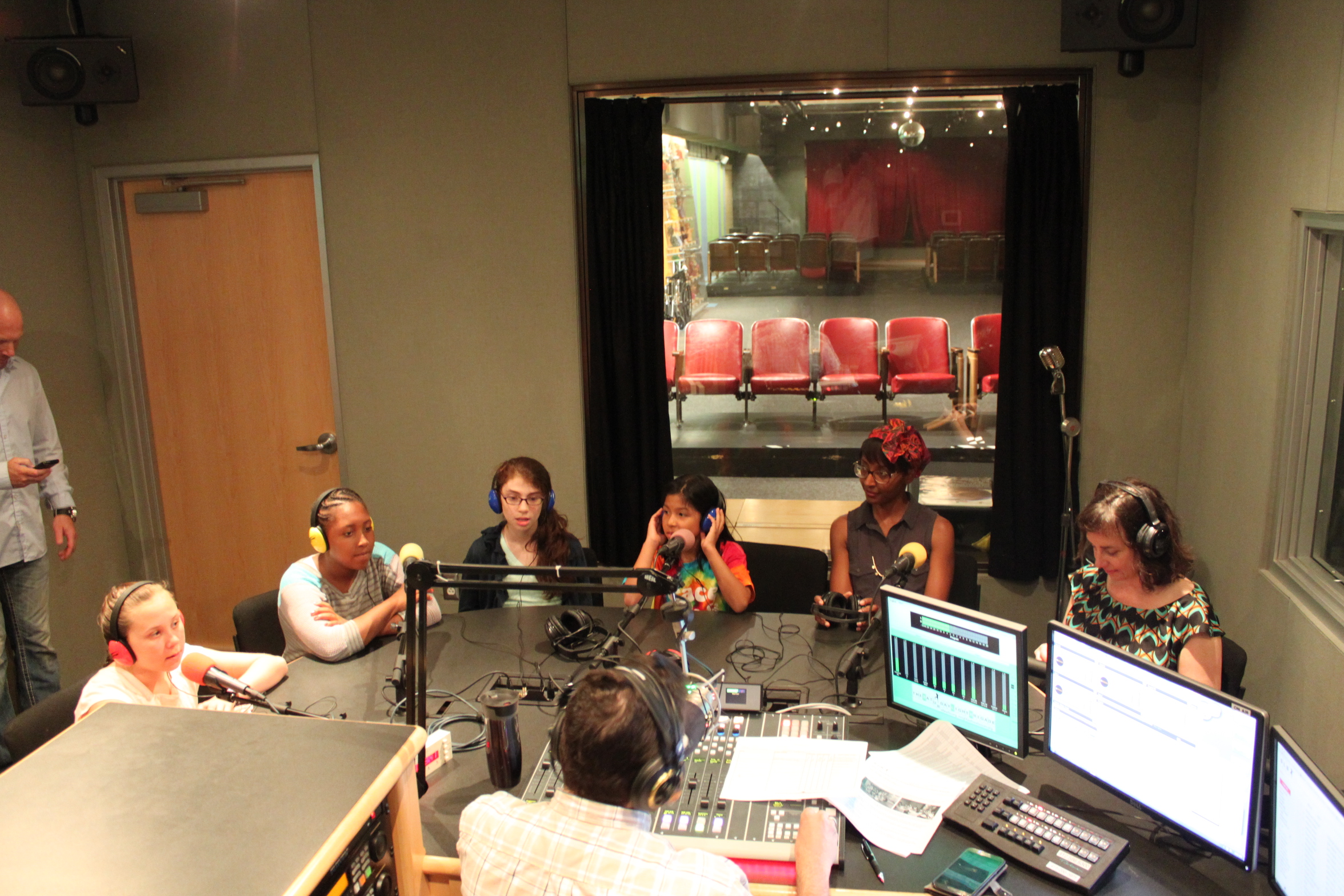 Four youth and two adults wearing headphones speaking into microphones being interviewed by an adult wearing headphones and sitting behind a microphone and a mixing board in a radio studio
