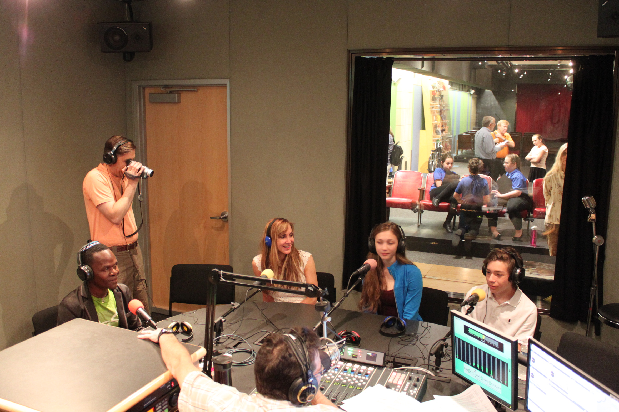 Four teens wearing headphones speaking into microphones being interviewed by an adult wearing headphones sitting behind a microphone and a mixing board as an adult standing in the background films with a camera in a radio studio