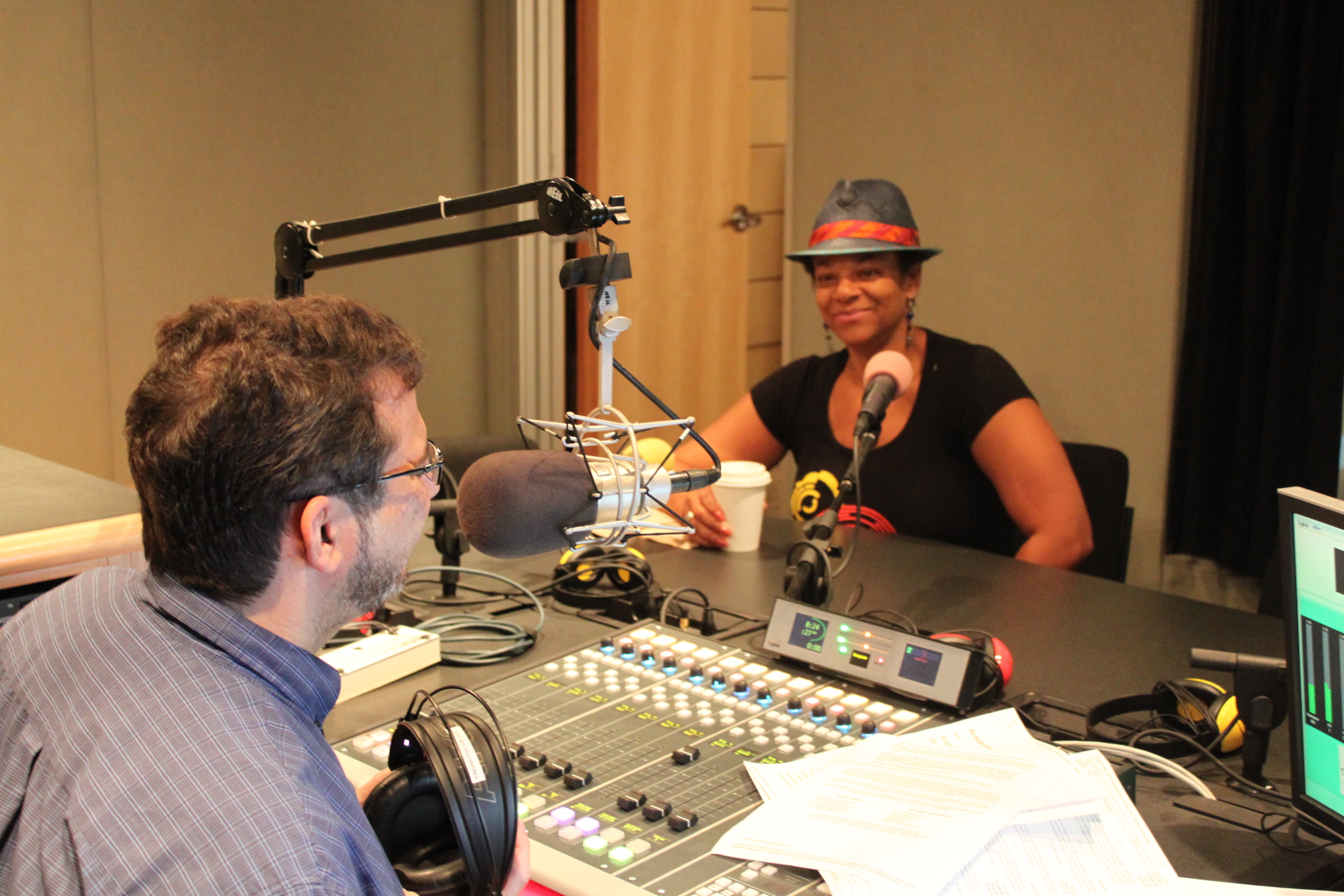 An adult sitting behind a microphone being interviewed by an adult speaking into a microphone sitting behind a mixing board in a radio studio