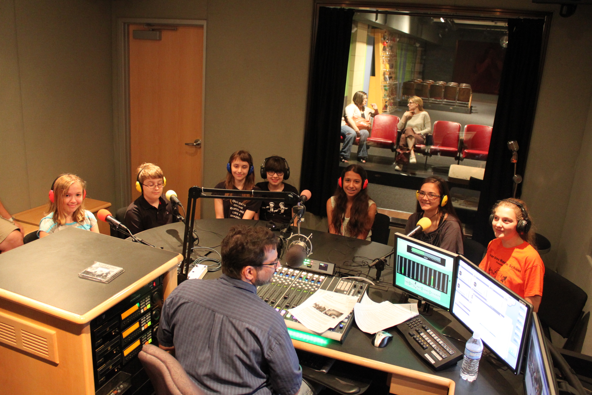 Seven youth wearing headphones speaking into microphones being interviewed by an adult wearing headphones sitting behind a microphone and a mixing board in a radio studio