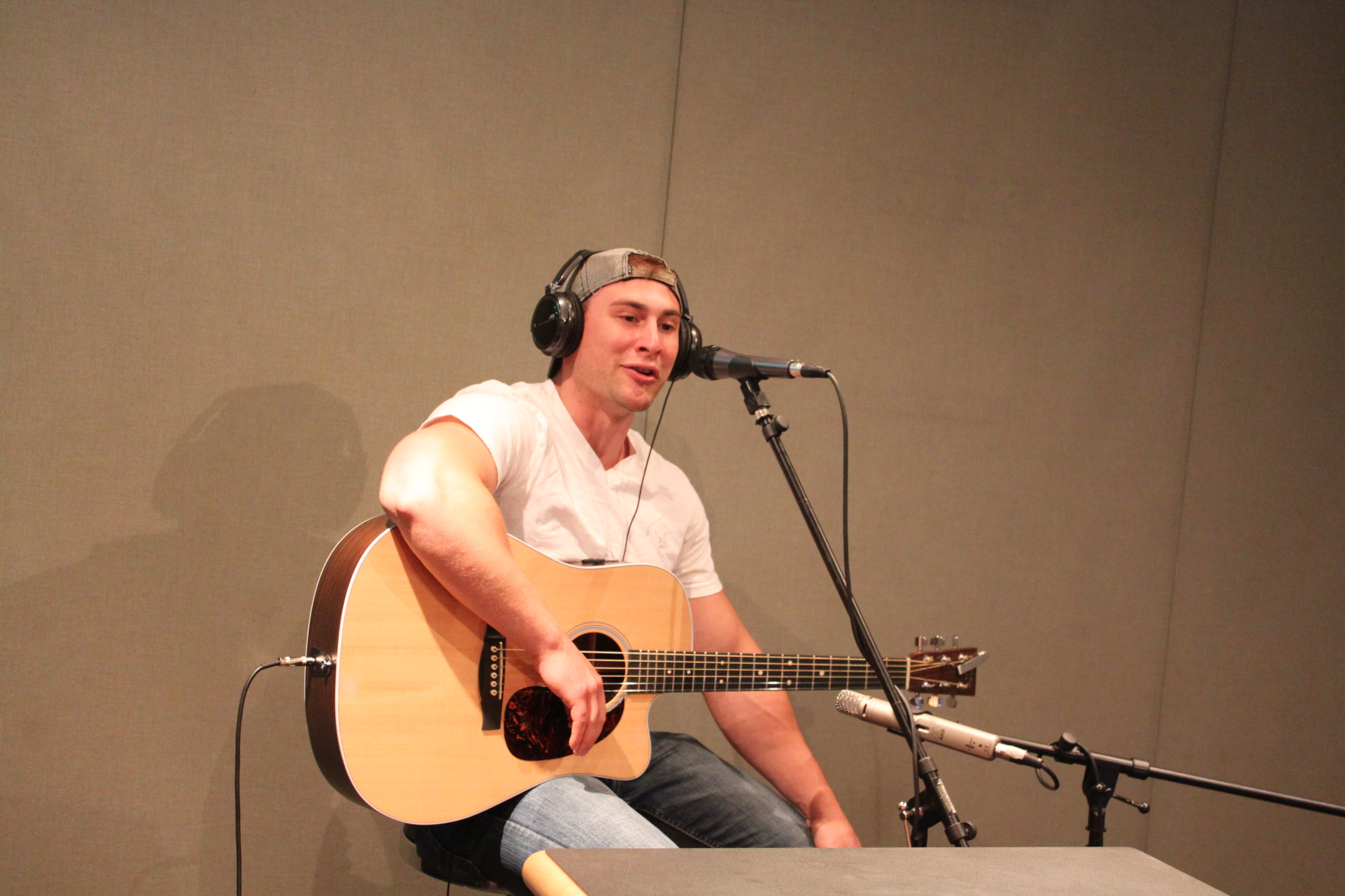 An adult wearing headphones singing into a microphone and playing acoustic guitar in a radio studio