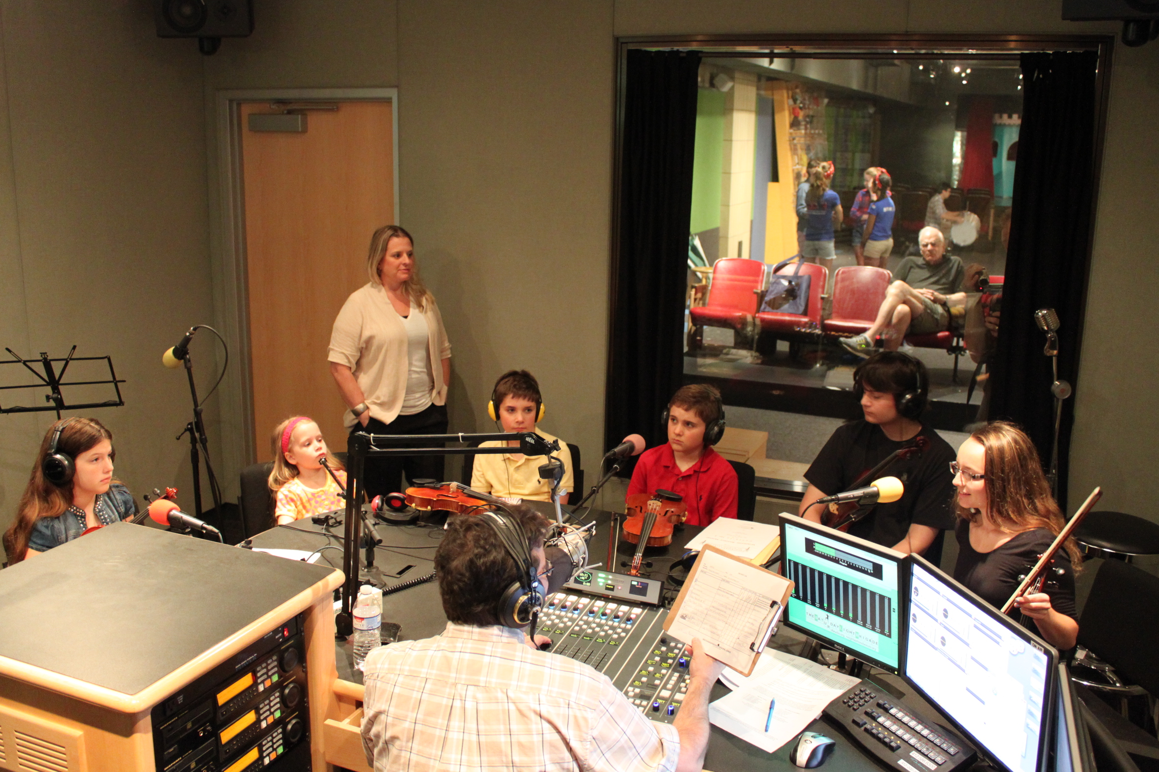 Five youth wearing headphones and one teen sitting behind microphones being interviewed by an adult wearing headphones sitting behind a microphone and a mixing board as another adult stands by and listens in a radio studio