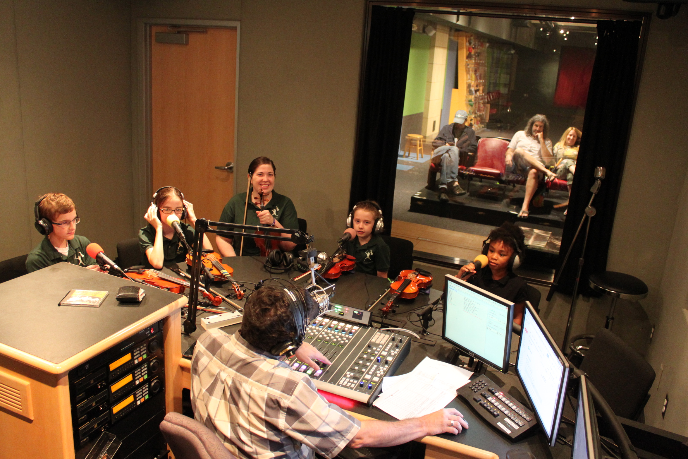 Four youth and one adult wearing headphones sitting behind microphones and violins being interviewed by an adult wearing headphones sitting behind a microphone and a mixing board in a radio studio