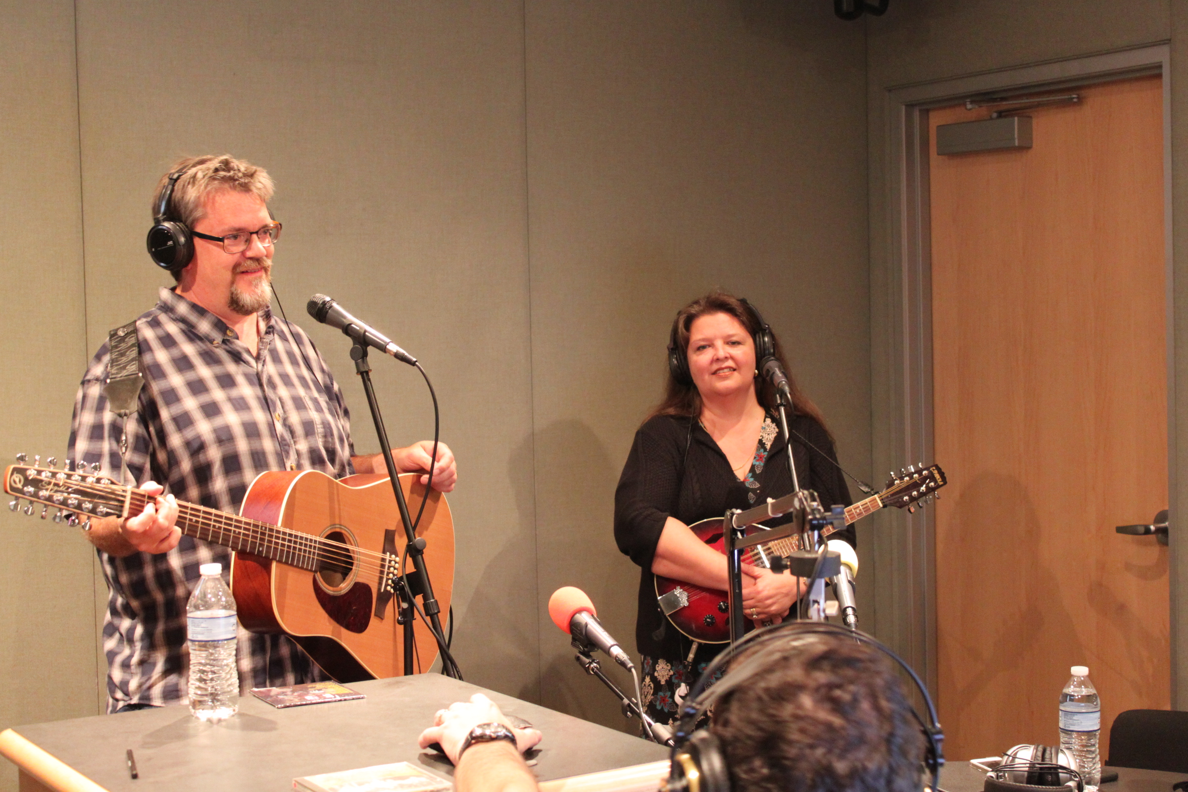 An adult wearing headphones holding a guitar and standing behind a microphone, and an adult wearing headphones holding a mandolin standing behind a microphone in a radio studio