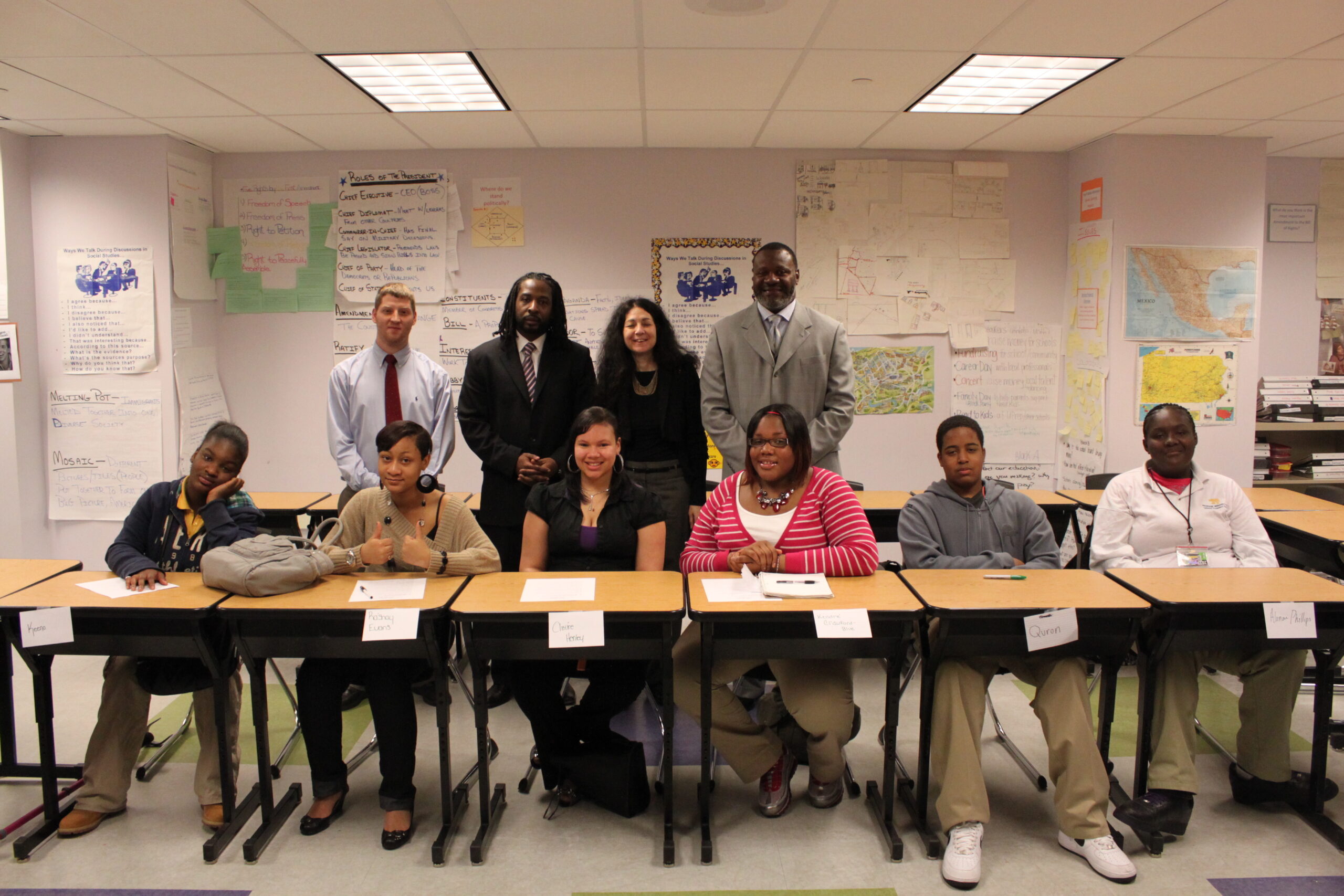 Six teens sitting at classroom desks and four adults standing behind them