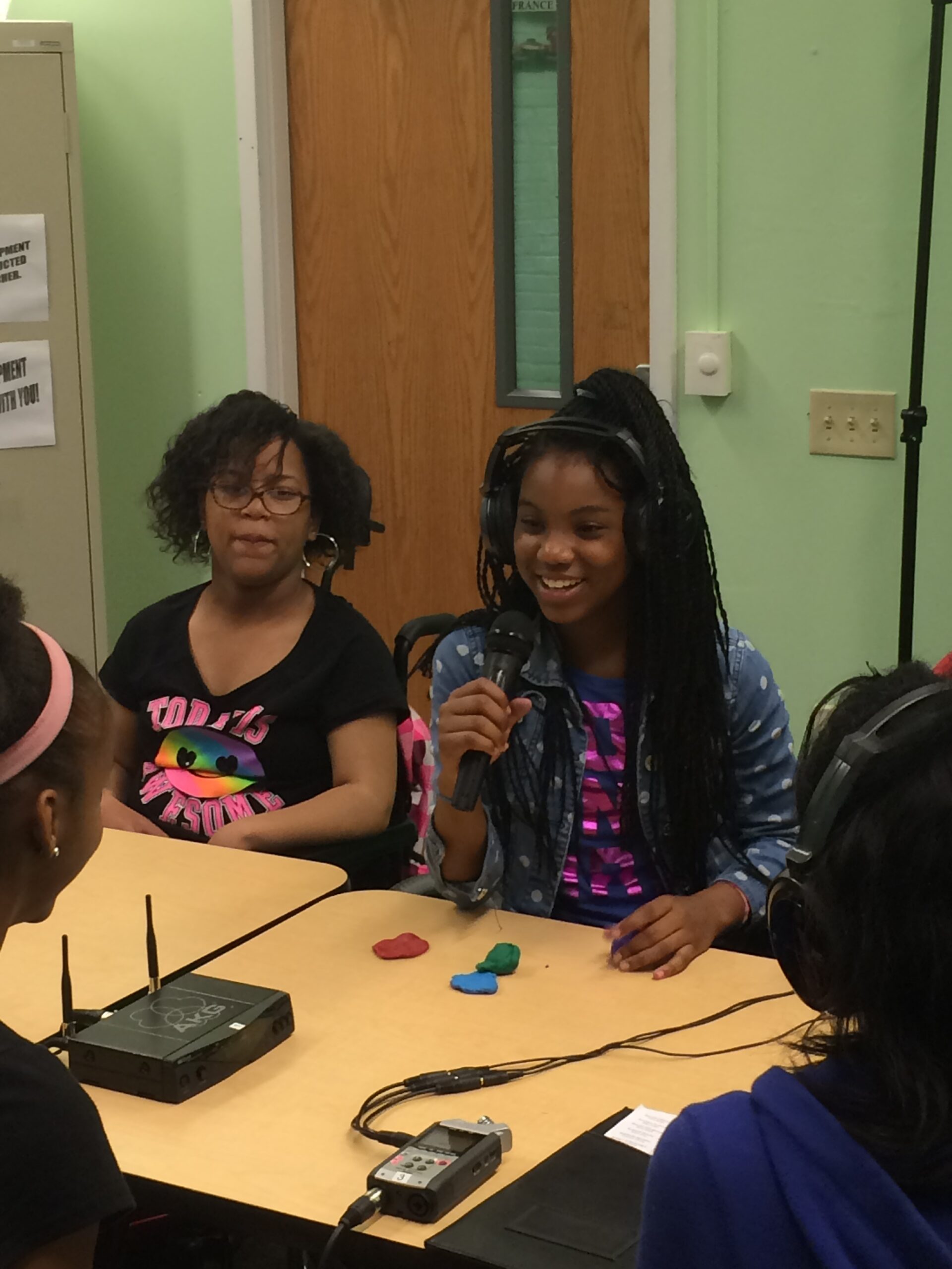 Four teens sitting around a table in a classroom, two wearing headphones, one holding and speaking into a microphone.