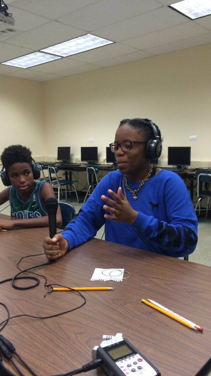 Two teens wearing headphones sitting around a brown table in a classroom, one holding a microphone