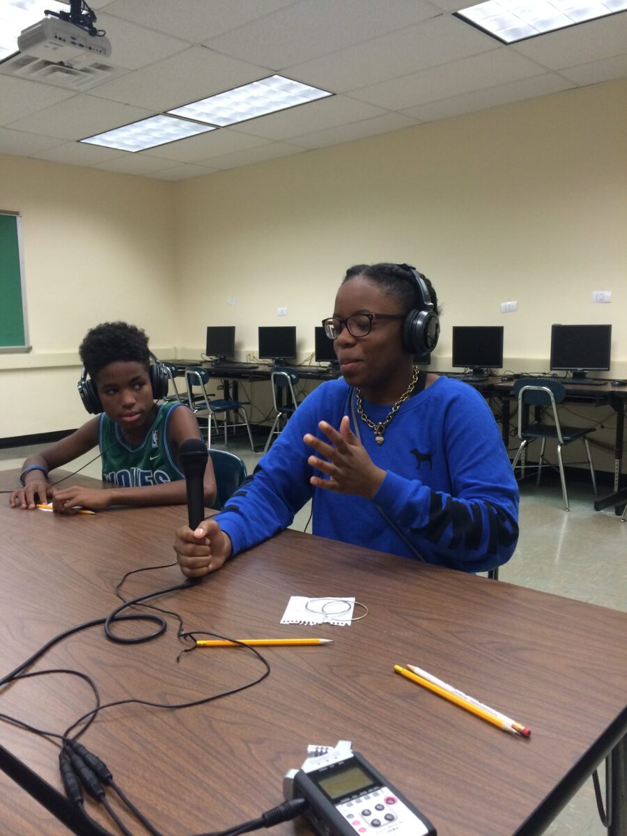IMG_0544 Two teens wearing headphones sitting around a brown table in a classroom, one holding a microphone