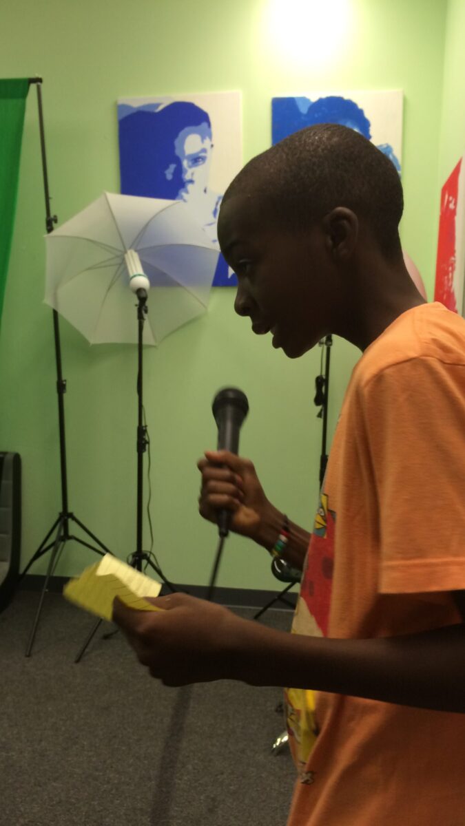 A youth looking at a sticky note and holding and speaking into a microphone in a green room